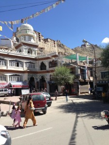 An Indian woman wearing a kurta walks her children across the busy corner of the main square in Leh in front of the mosque, with Leh Palace in the background and Tsemo Fort even further up.